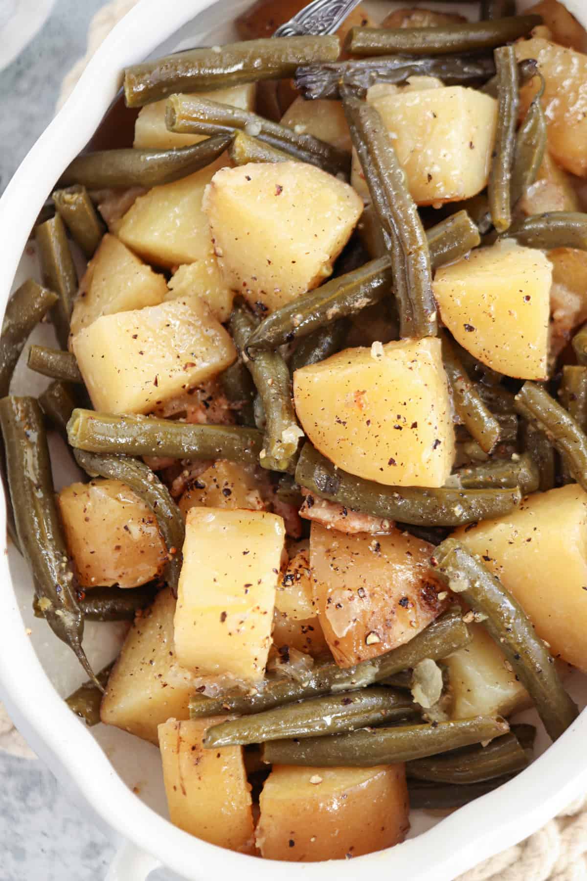 A close-up of cooked green beans and cubed potatoes seasoned with black pepper, reminiscent of the flavors in a chicken potato broccoli casserole, served in a white dish.
