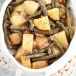 A close-up of a white bowl filled with cooked potatoes and green beans, seasoned with black pepper and spices, reminiscent of a chicken potato broccoli casserole. The vegetables are cut into chunks and appear soft and well-cooked.