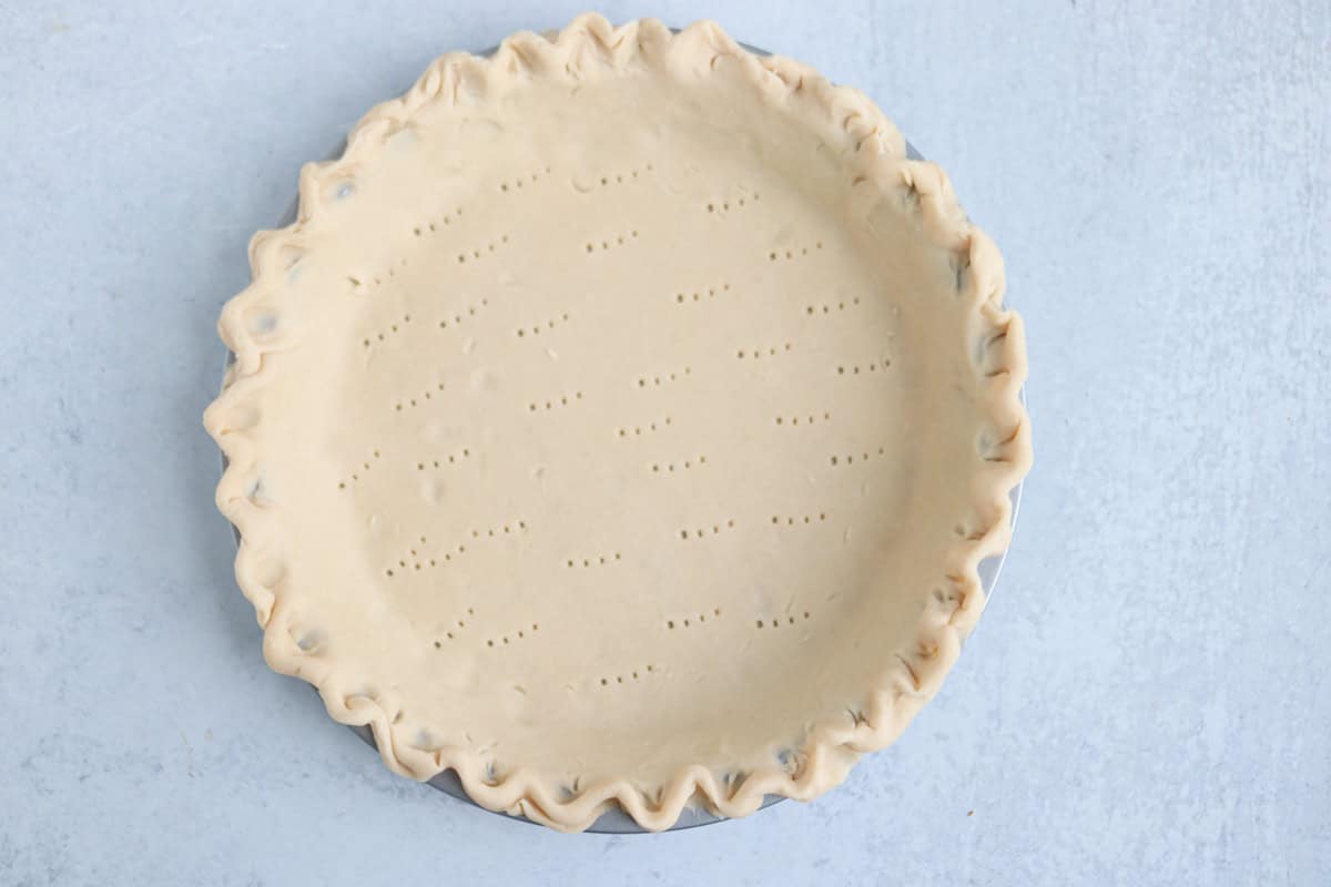 A pie dish filled with unbaked pie crust, edges crimped, and the base pricked with a fork, ready to be filled for a hearty chicken potato broccoli casserole, sits on a light gray surface.