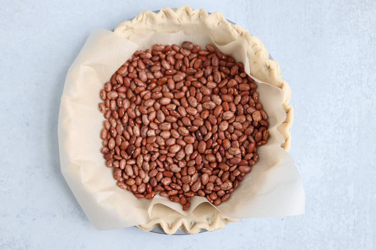 A pie crust lined with parchment paper and filled with dry beans, used as pie weights, sits on a light gray surface. Perfectly crimped edges make it ideal for blind baking before creating dishes like chicken potato broccoli casserole.