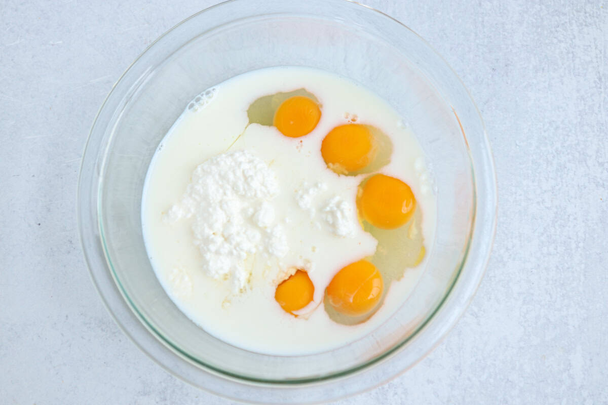 A glass bowl containing five cracked eggs, milk, and a dollop of cottage cheese on a light gray surface—perfect ingredients for starting a chicken potato broccoli casserole.