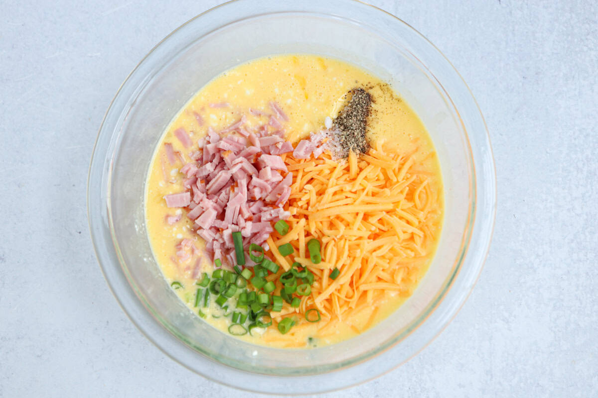 A glass bowl filled with whisked eggs, chopped ham, shredded cheddar cheese, sliced green onions, black pepper, and salt—ingredients that could inspire a delicious chicken potato broccoli casserole—sits on a light gray surface.