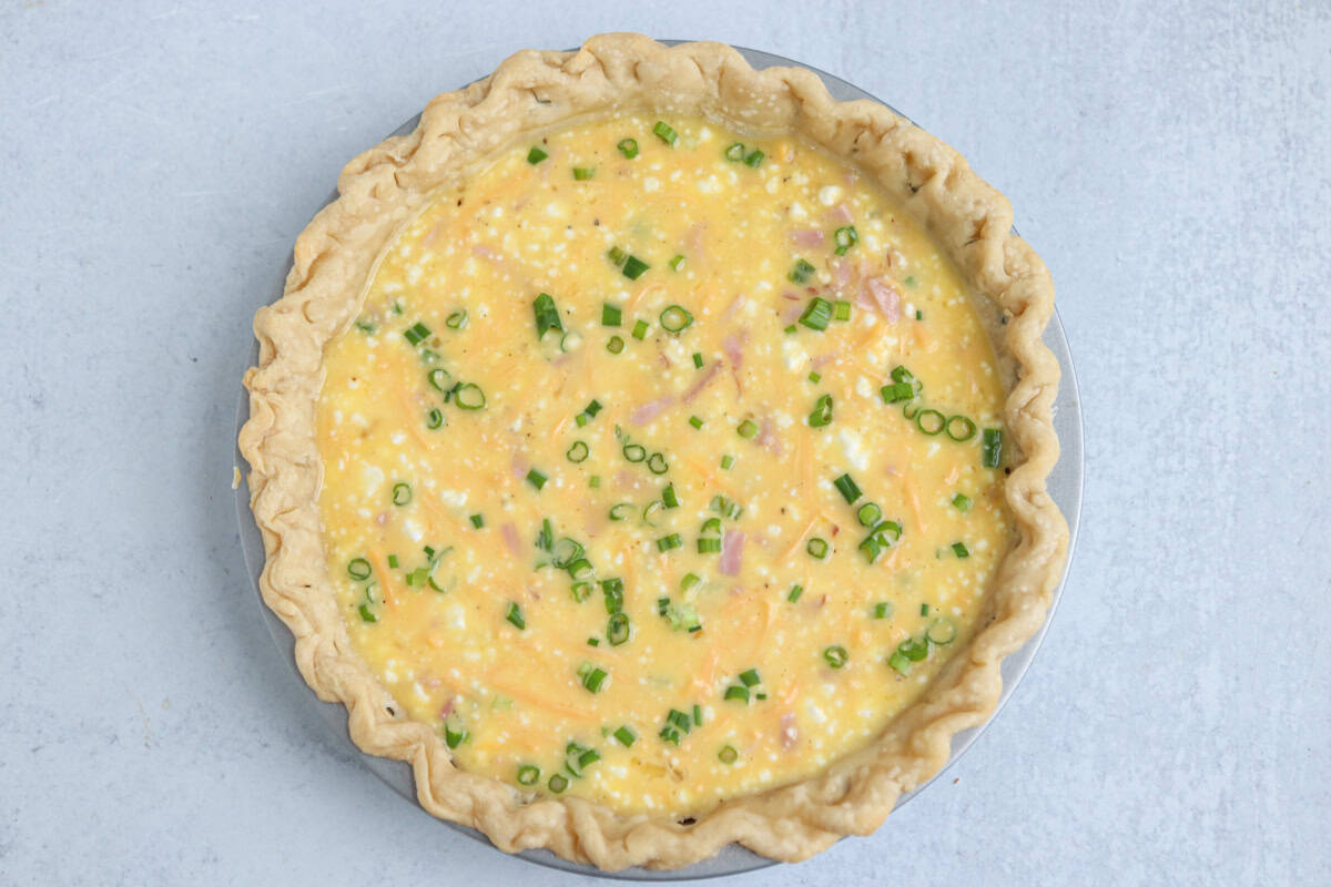 Unbaked quiche in a pie crust, filled with an egg mixture, diced ham, shredded cheese, and chopped green onions—perfect alongside a hearty chicken potato broccoli casserole—on a light gray surface.