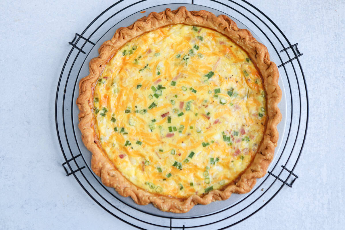 A freshly baked quiche with a golden, flaky crust sits on a black wire cooling rack. Reminiscent of a chicken potato broccoli casserole, the filling appears to include cheese, green onions, and bits of ham. The background is a light gray surface.