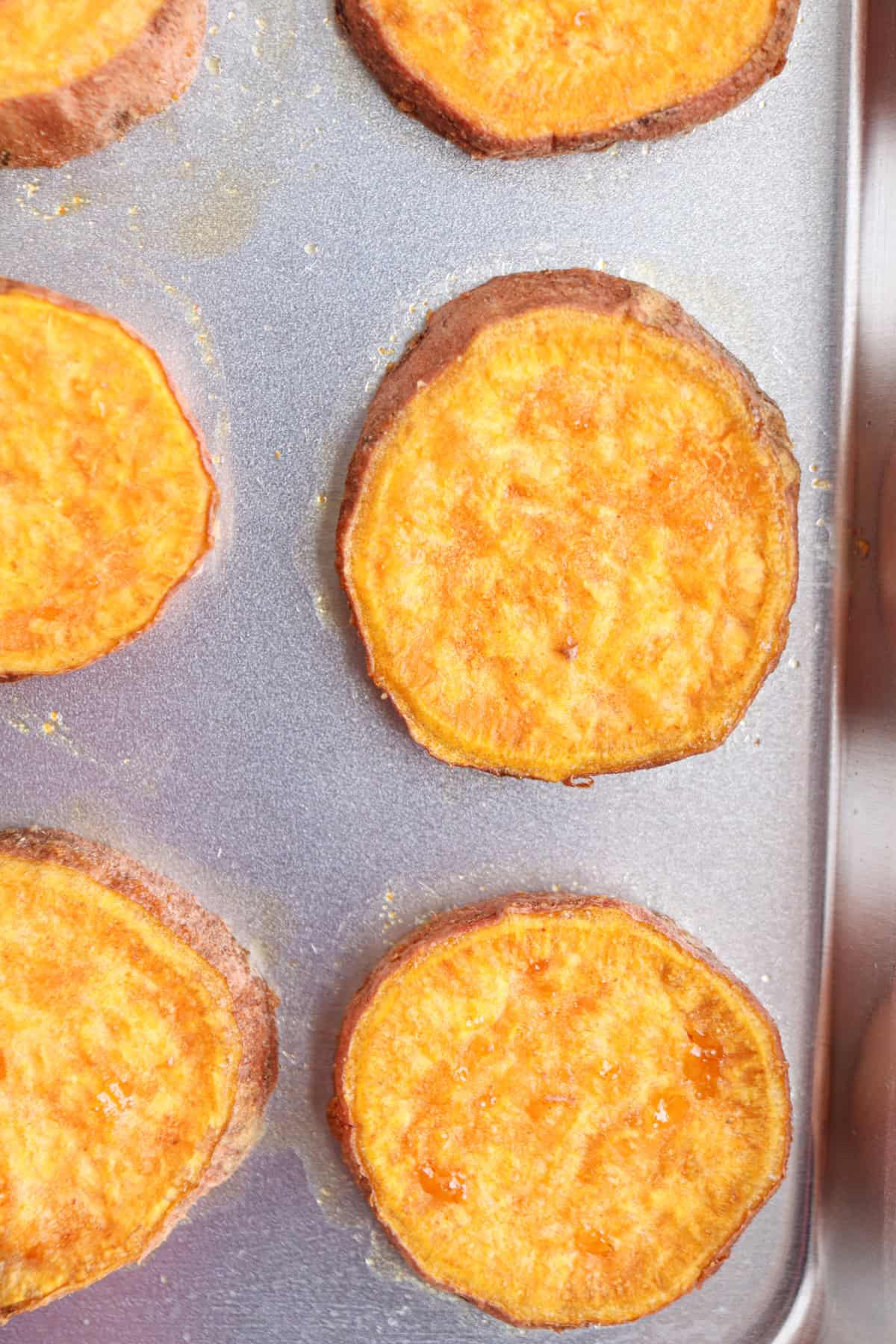 Roasted sweet potato slices are arranged on a metal baking sheet. The rounds are golden-orange in color, with slightly crispy, browned edges.