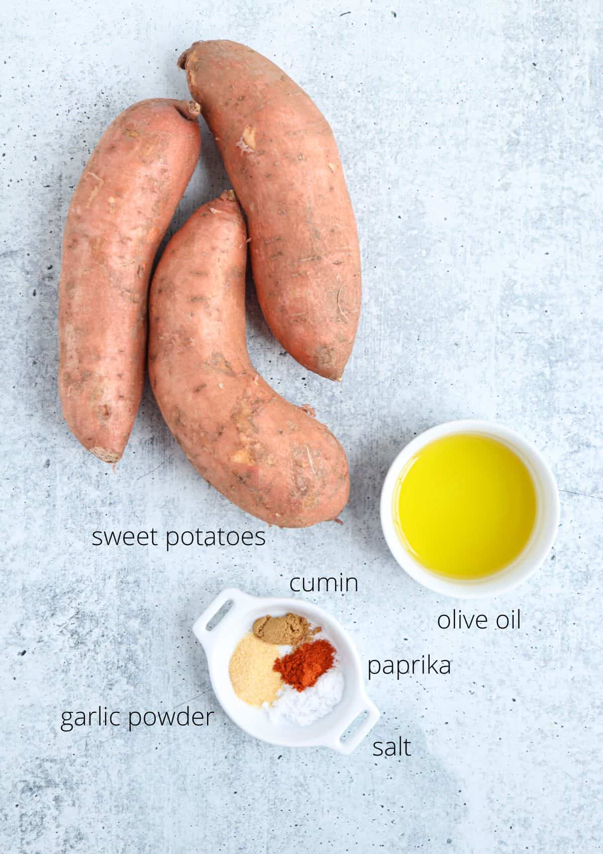Three whole sweet potatoes, a bowl of olive oil, and a small dish containing garlic powder, cumin, paprika, and salt are arranged on a light gray surface—perfect ingredients to inspire your next chicken potato broccoli casserole. Each item is labeled.