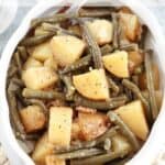 A white serving dish filled with slow cooker potatoes and green beans, seasoned with black pepper. A serving spoon is partially visible in the food. Text at the top reads, Slow Cooker Green Beans and Potatoes.