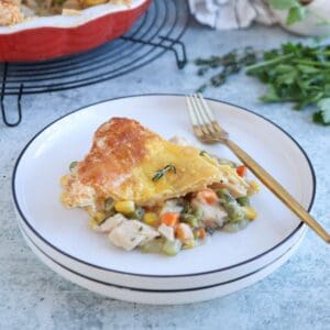 A slice of chicken pot pie with golden crust, filled with chicken, carrots, corn, and green beans—reminiscent of a classic chicken potato broccoli casserole—served on a white plate with a gold fork. Fresh herbs and the pie dish are in the background.