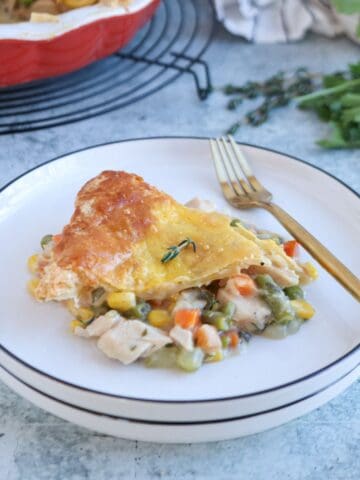 A serving of chicken pot pie with a golden, flaky crust sits on a white plate with a gold fork. Similar to a chicken potato broccoli casserole, the filling includes chicken, carrots, corn, and green beans. Fresh herbs are visible in the background.
