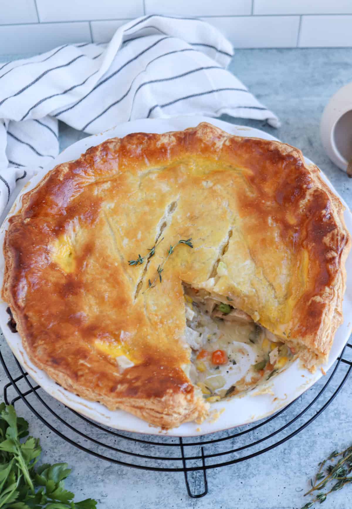 A golden-brown chicken and potato pot pie with a flaky crust sits on a wire rack. A large slice has been removed, revealing a creamy vegetable and meat filling—reminiscent of a comforting casserole. Fresh herbs and a striped towel are nearby.