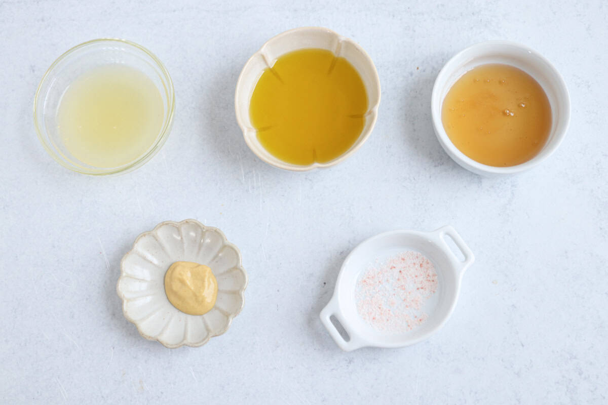 Five small dishes on a white surface, each holding an ingredient perfect for a chicken potato broccoli casserole: lemon juice, olive oil, honey, Dijon mustard, and a small dish of pinkish salt.