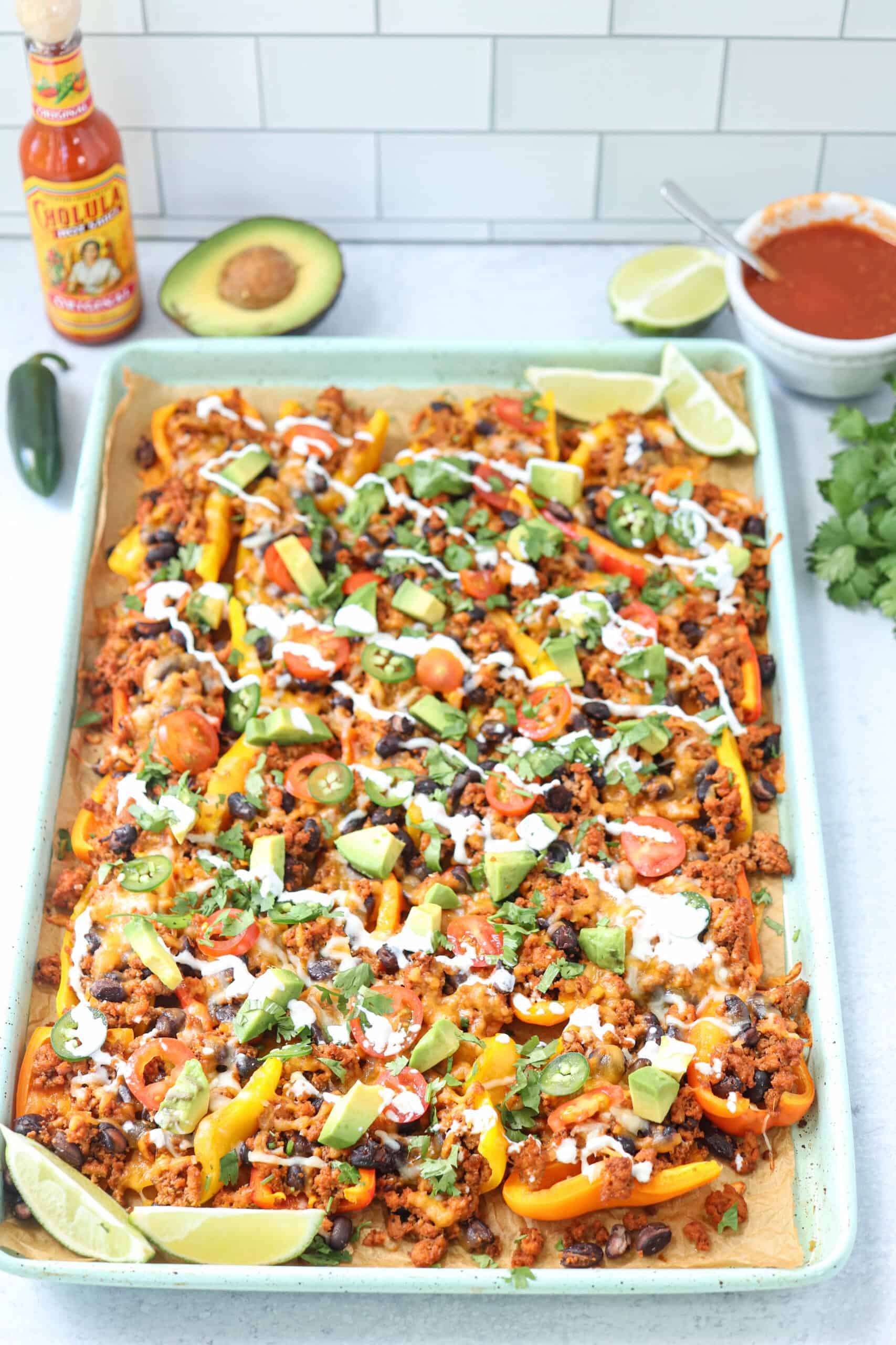 A baking sheet filled with colorful nachos topped with ground meat, black beans, avocado, jalapeños, tomatoes, and drizzled with sour cream. Lime wedges, half an avocado, salsa, and hot sauce are nearby on a white counter.