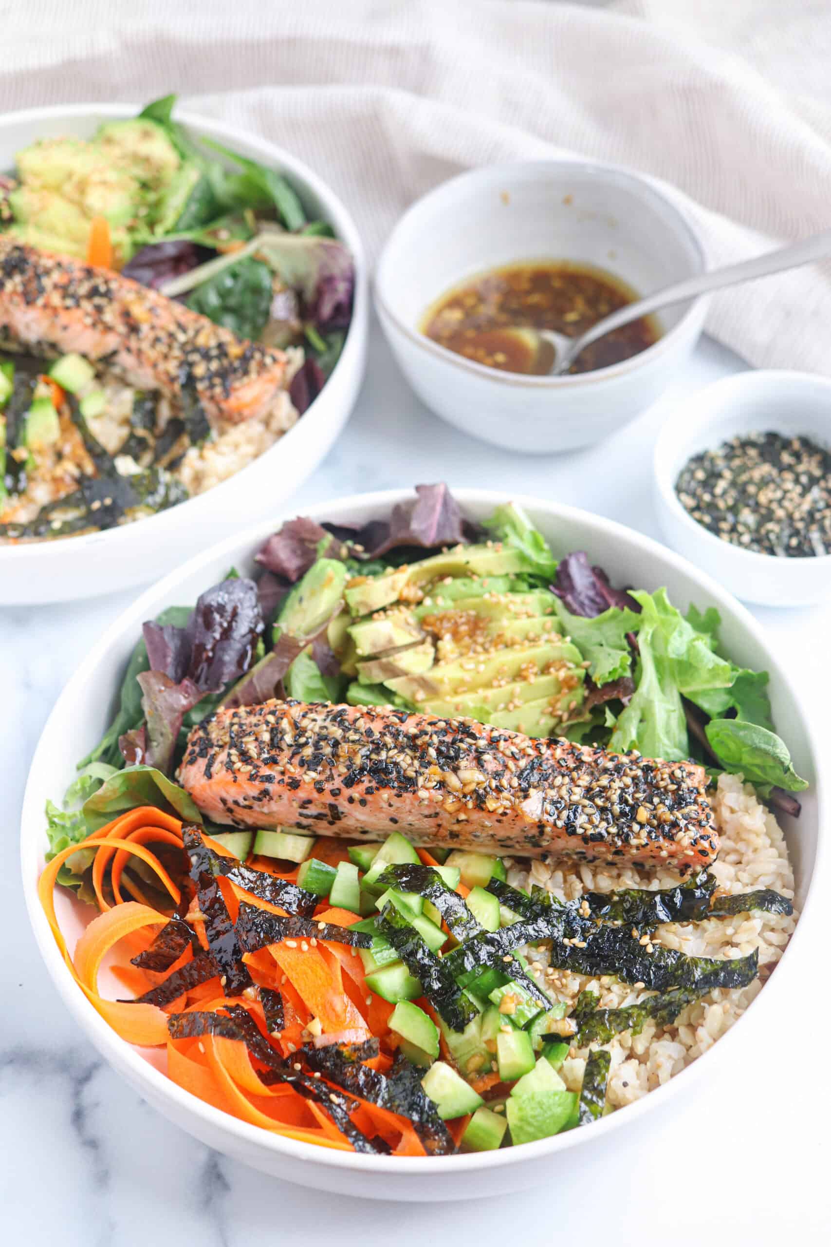 Two bowls of salmon poke with sesame-crusted salmon, sliced avocado, greens, julienned carrots, cucumber, seaweed strips, and rice. A small bowl of sauce and a dish of sesame seeds sit nearby.