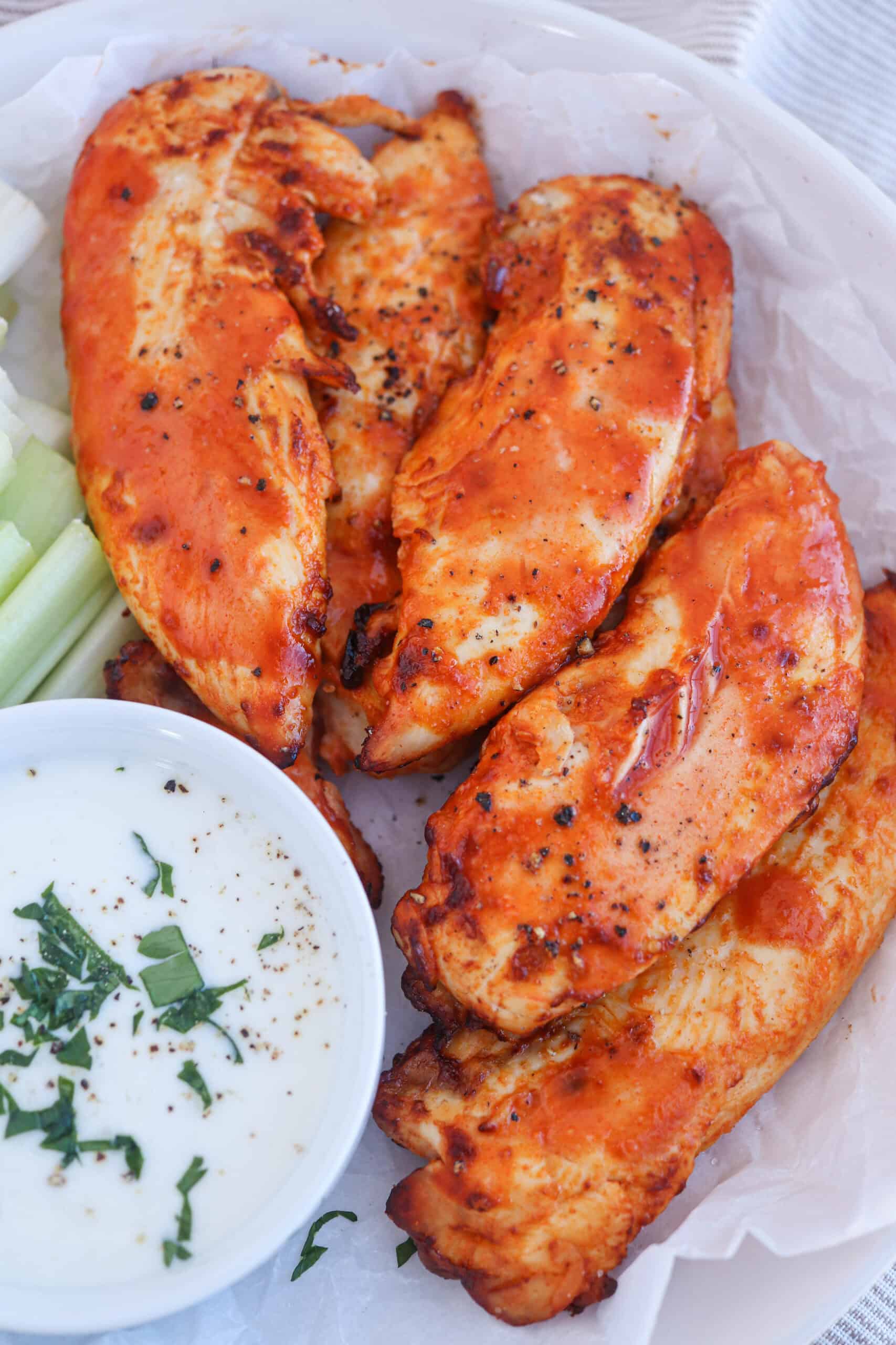 A plate of buffalo chicken tenders with a side of celery sticks and a small bowl of ranch dipping sauce garnished with chopped herbs.