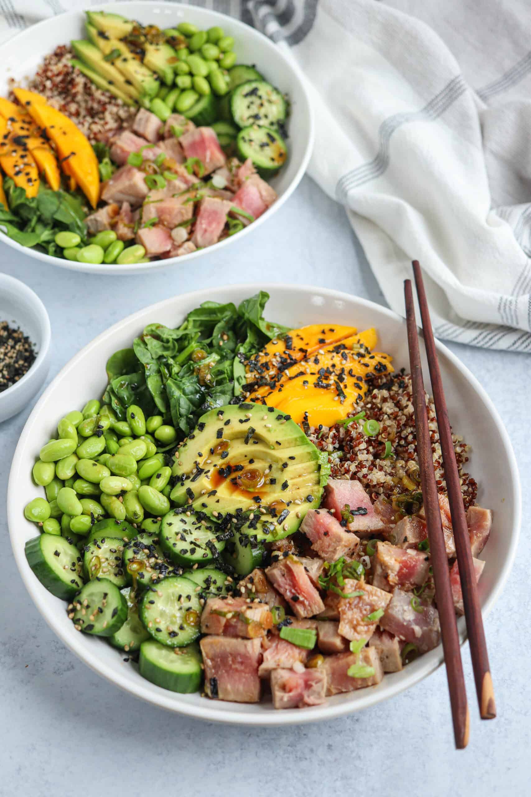 A colorful poke bowl filled with cubed seared tuna, sliced avocado, edamame, cucumber, quinoa, spinach, mango, and black sesame seeds, with chopsticks resting on the bowl and a napkin nearby.