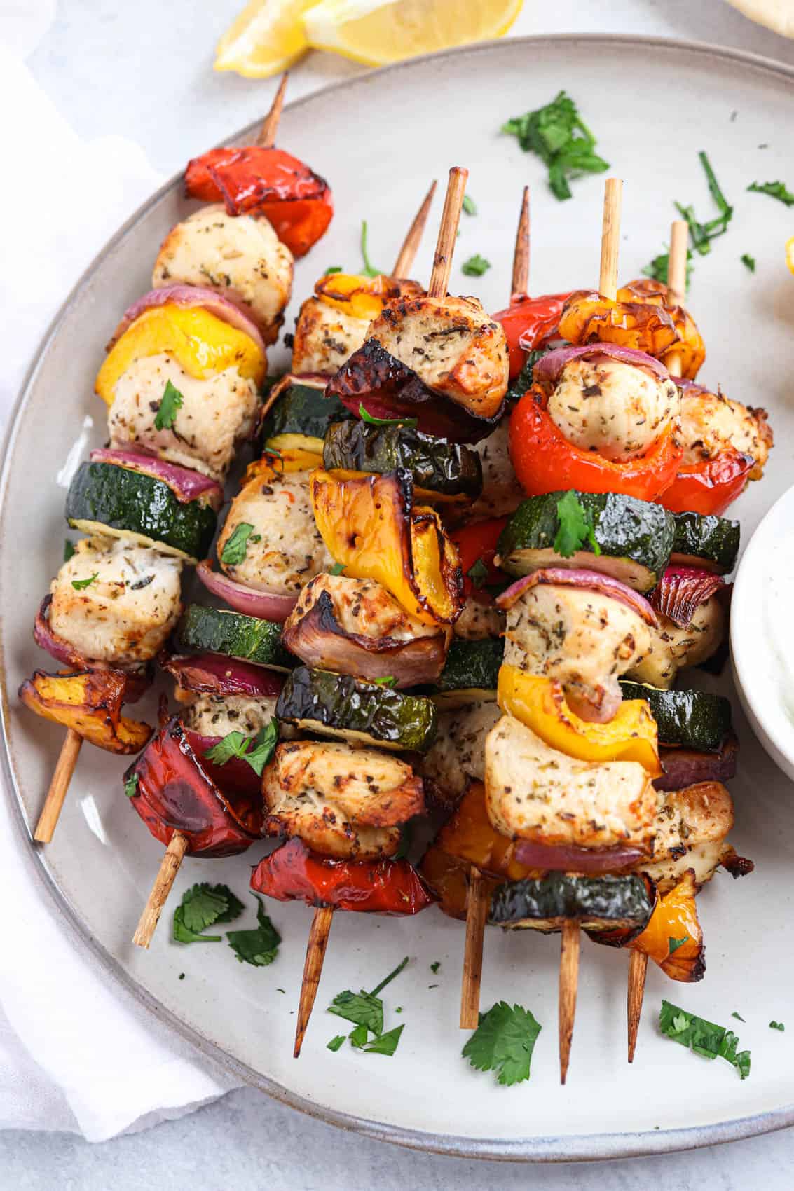 Grilled chicken and vegetable skewers with red onion, zucchini, and bell peppers, garnished with chopped herbs, served on a plate with lemon wedges in the background.