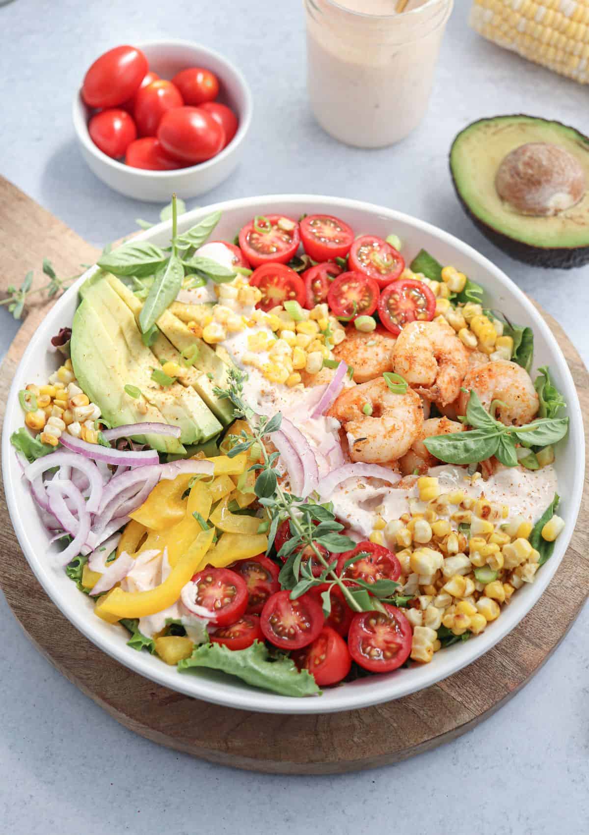 A colorful salad bowl with shrimp, sliced avocado, cherry tomatoes, yellow bell pepper, red onion, corn, greens, and herbs. A jar of dressing, avocado half, and a bowl of cherry tomatoes are in the background.