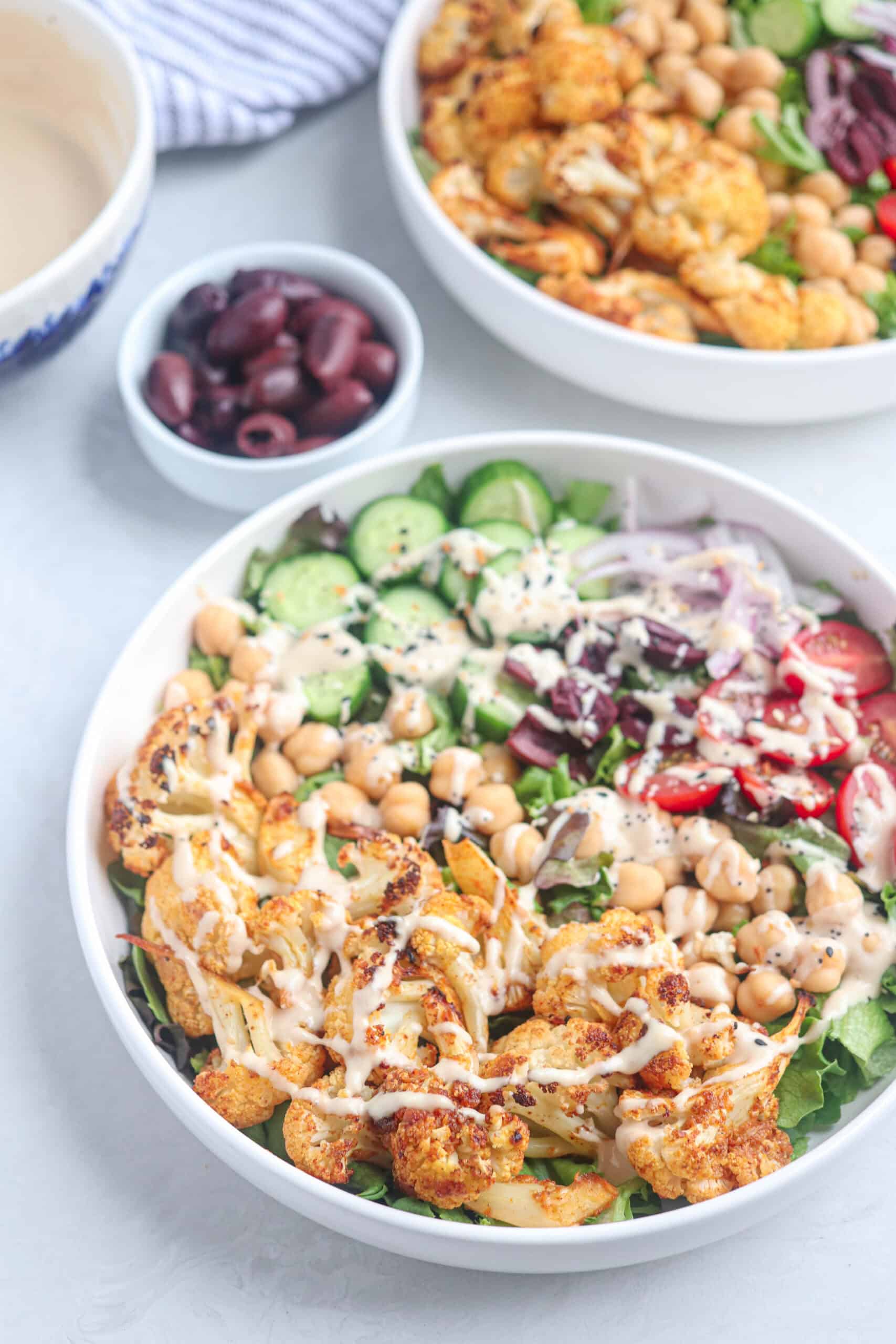 A bowl of salad topped with roasted cauliflower, chickpeas, cucumber, red onion, tomatoes, olives, and a drizzle of creamy dressing. A small bowl of olives and another salad bowl are in the background.