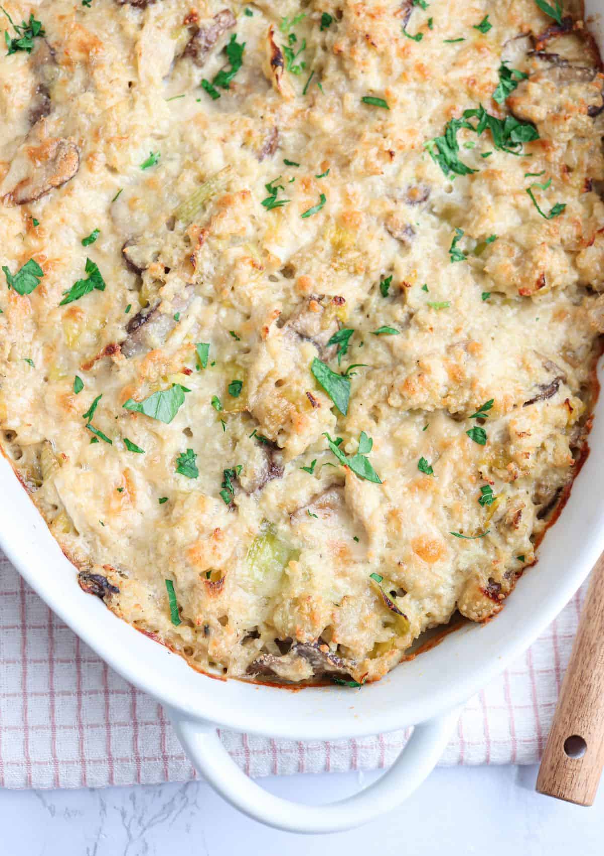 A close-up of a baked casserole topped with melted cheese and garnished with fresh chopped parsley in a white dish, sitting on a folded checked kitchen towel.