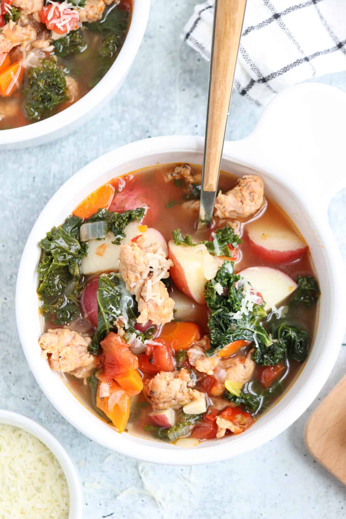A white bowl filled with sausage, kale, carrots, potatoes, and tomatoes in a hearty soup. A spoon is in the bowl, and grated cheese and a checkered napkin are nearby. Another bowl of soup is partially visible.