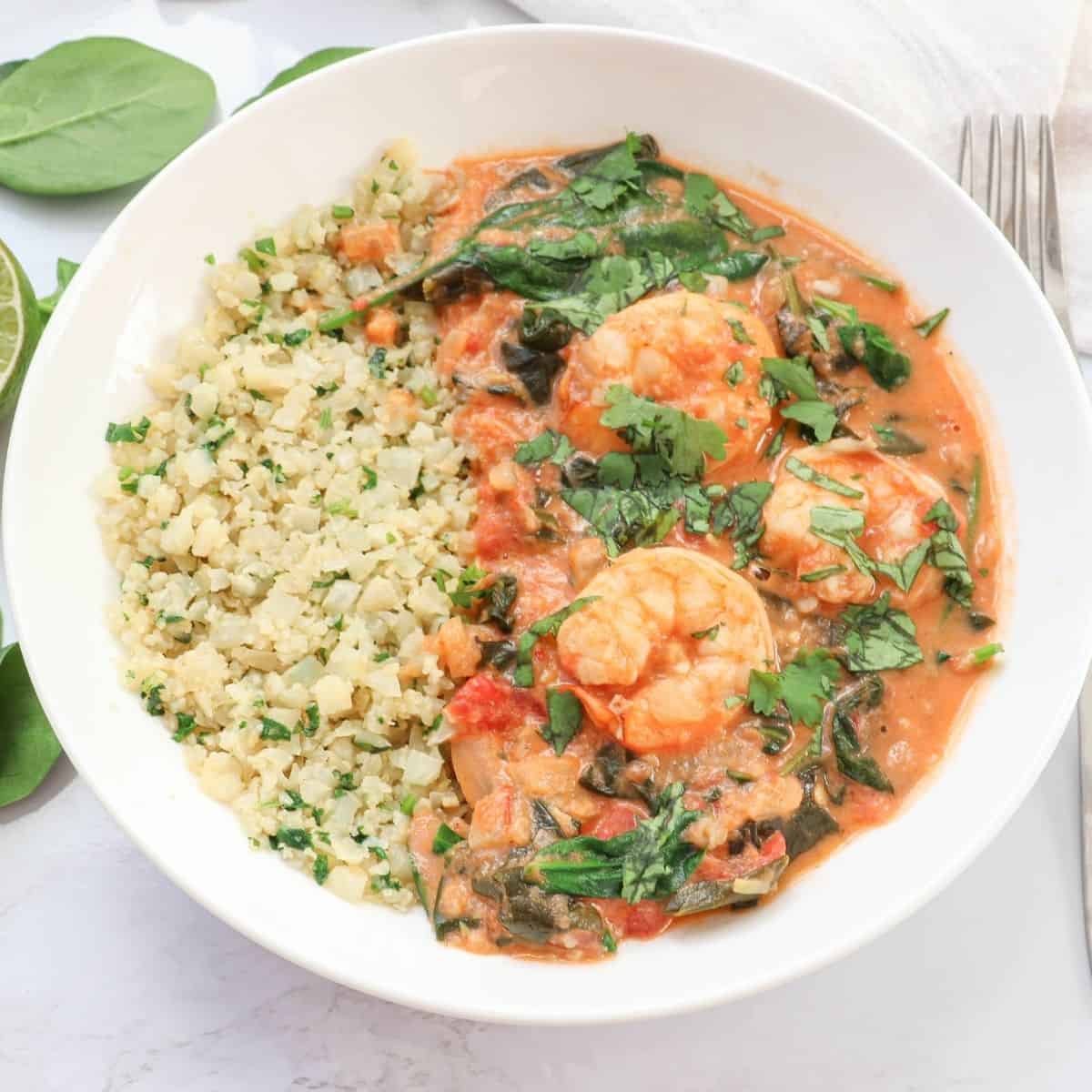 A white bowl filled with shrimp in a creamy tomato sauce with spinach, garnished with chopped herbs, served alongside a portion of cauliflower rice. A fork and spinach leaves are nearby.
