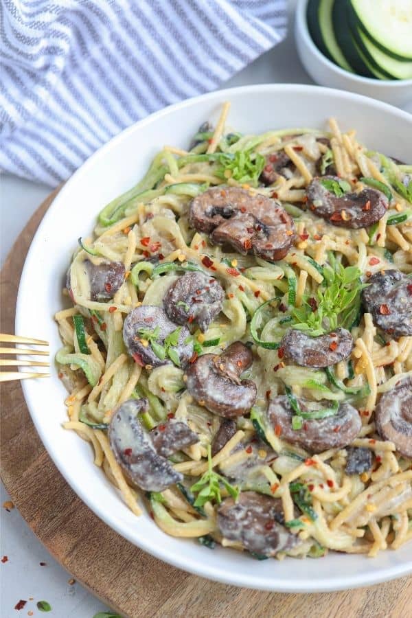 A white bowl filled with creamy pasta topped with sautéed mushrooms, fresh herbs, and red pepper flakes, set on a wooden board with a gold fork and a bowl of sliced cucumbers in the background.