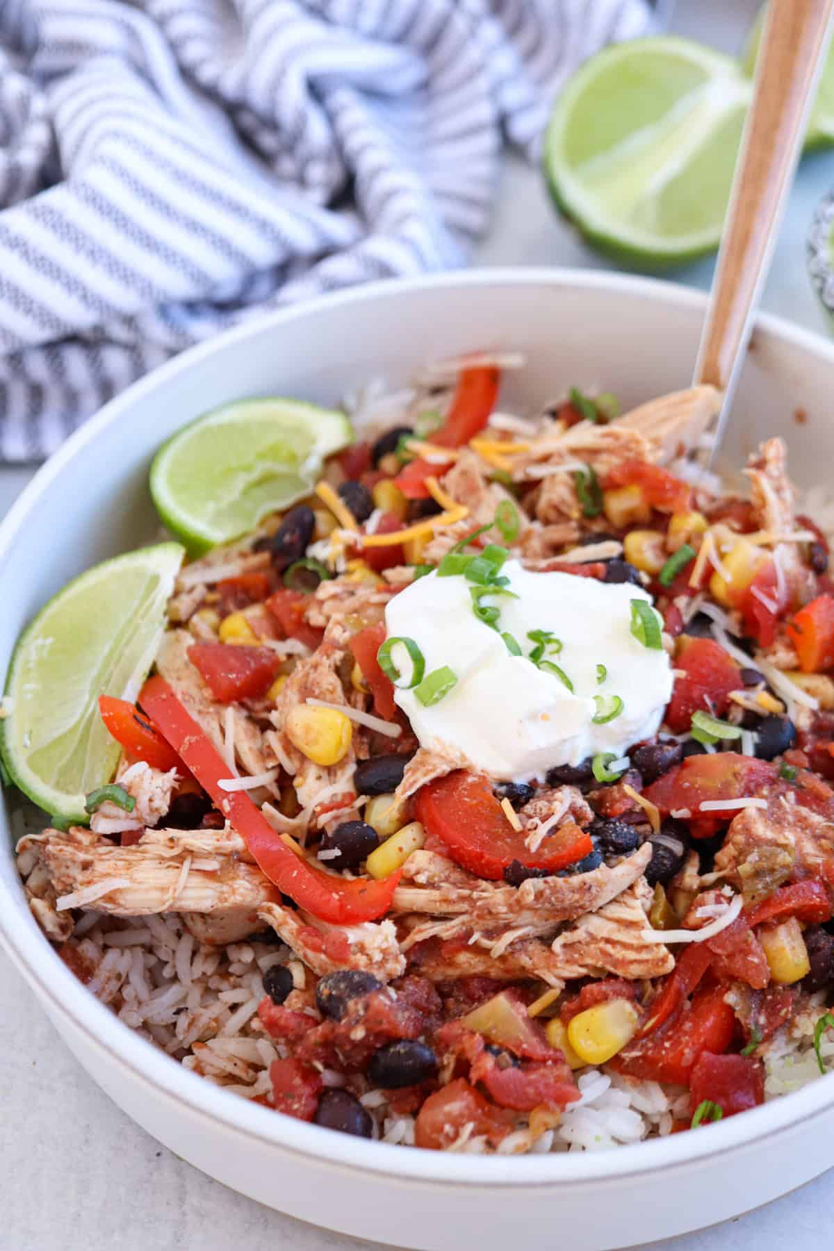 A bowl of rice topped with shredded chicken, black beans, corn, red bell peppers, diced tomatoes, shredded cheese, sour cream, chopped green onions, and lime wedges. A striped cloth and lime halves are in the background.