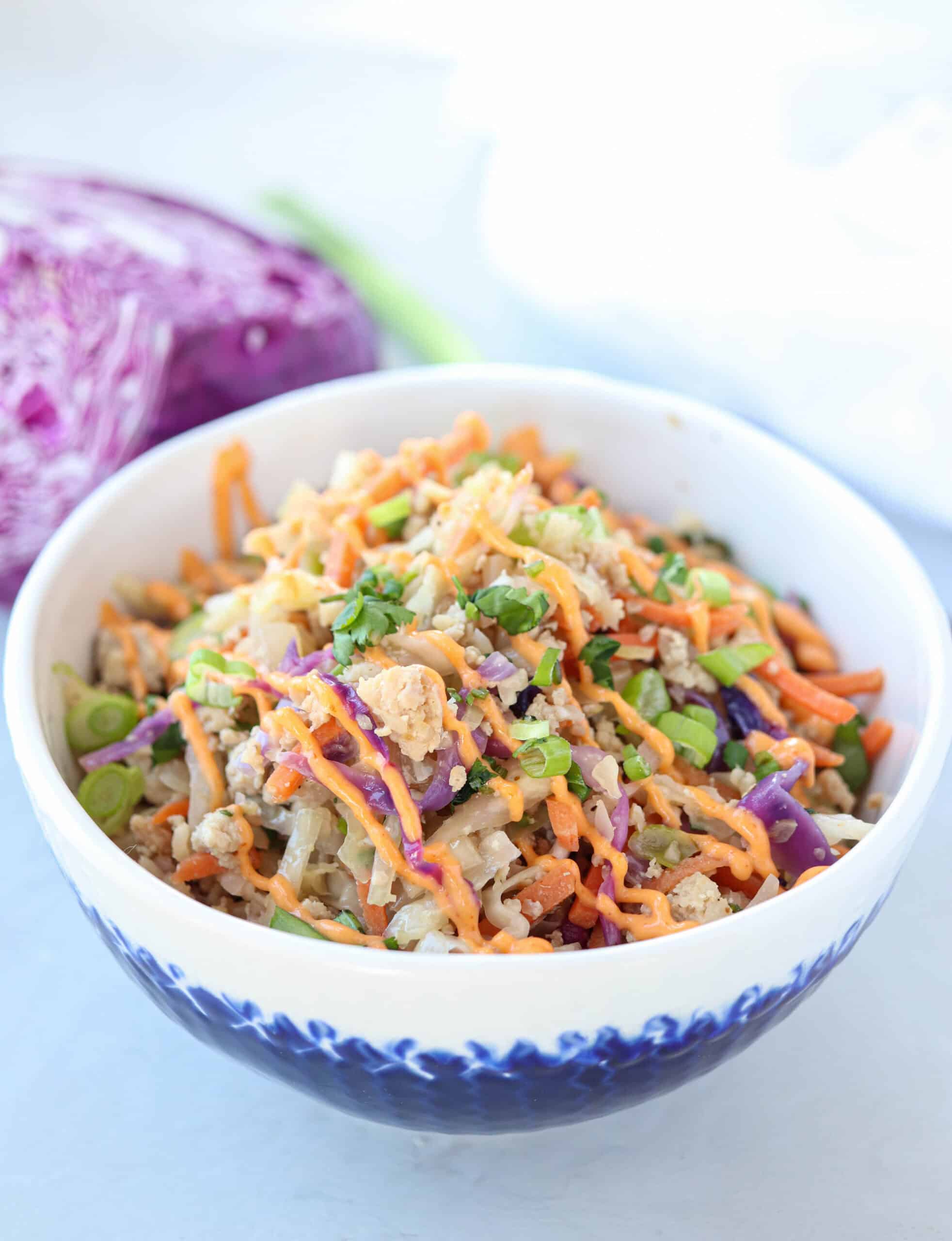A bowl filled with a colorful salad containing shredded carrots, cabbage, green onions, and a drizzle of orange dressing, with purple cabbage and a green onion in the background.