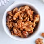 A white bowl filled with glazed candied walnuts, with a few walnuts scattered outside the bowl on a light surface—perfect as a sweet snack or a crunchy topping for your favorite chicken potato broccoli casserole.