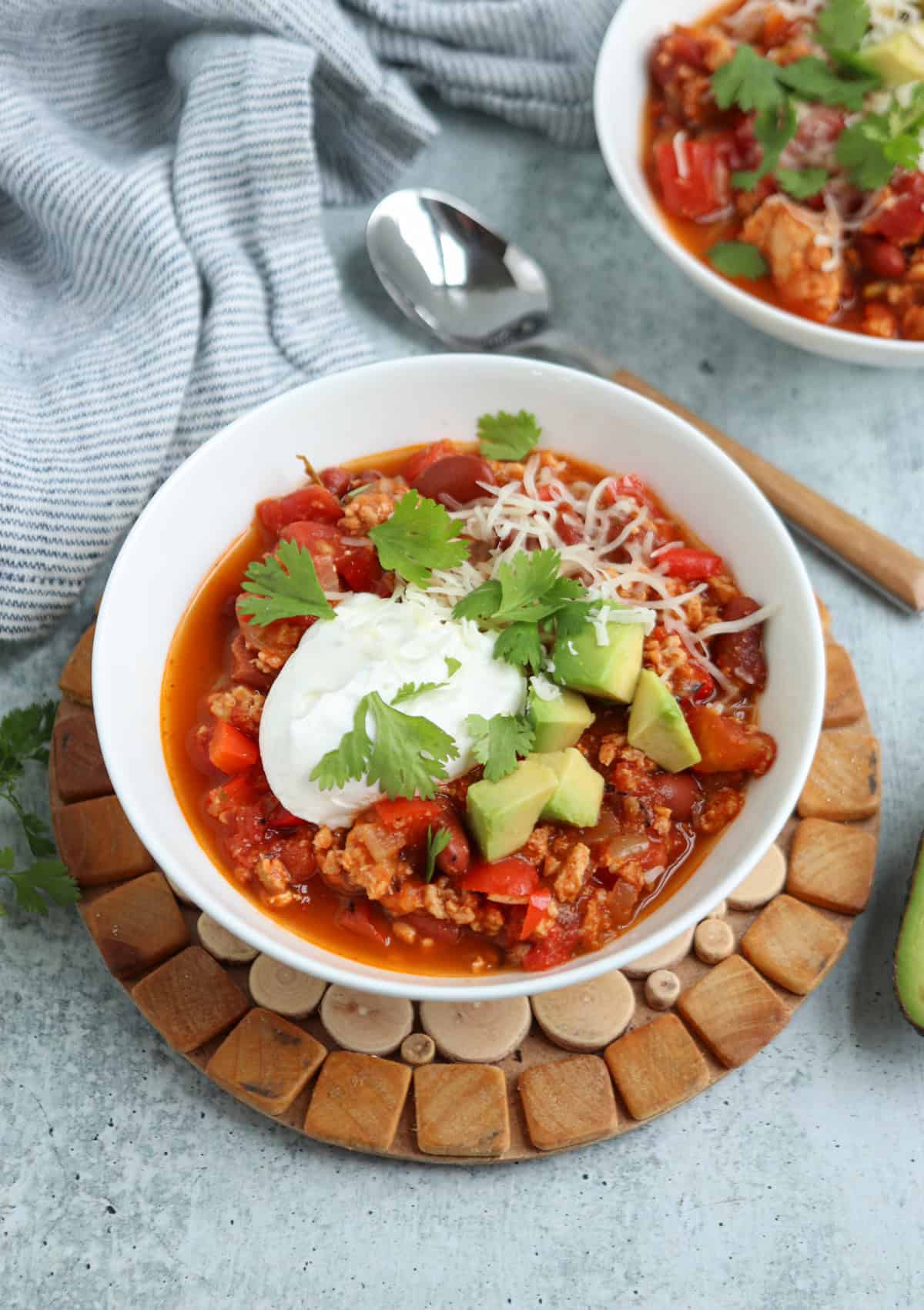 A bowl of chili topped with sour cream, shredded cheese, avocado chunks, and cilantro sits on a round wooden trivet. A spoon and another bowl of chili are in the background on a gray surface.