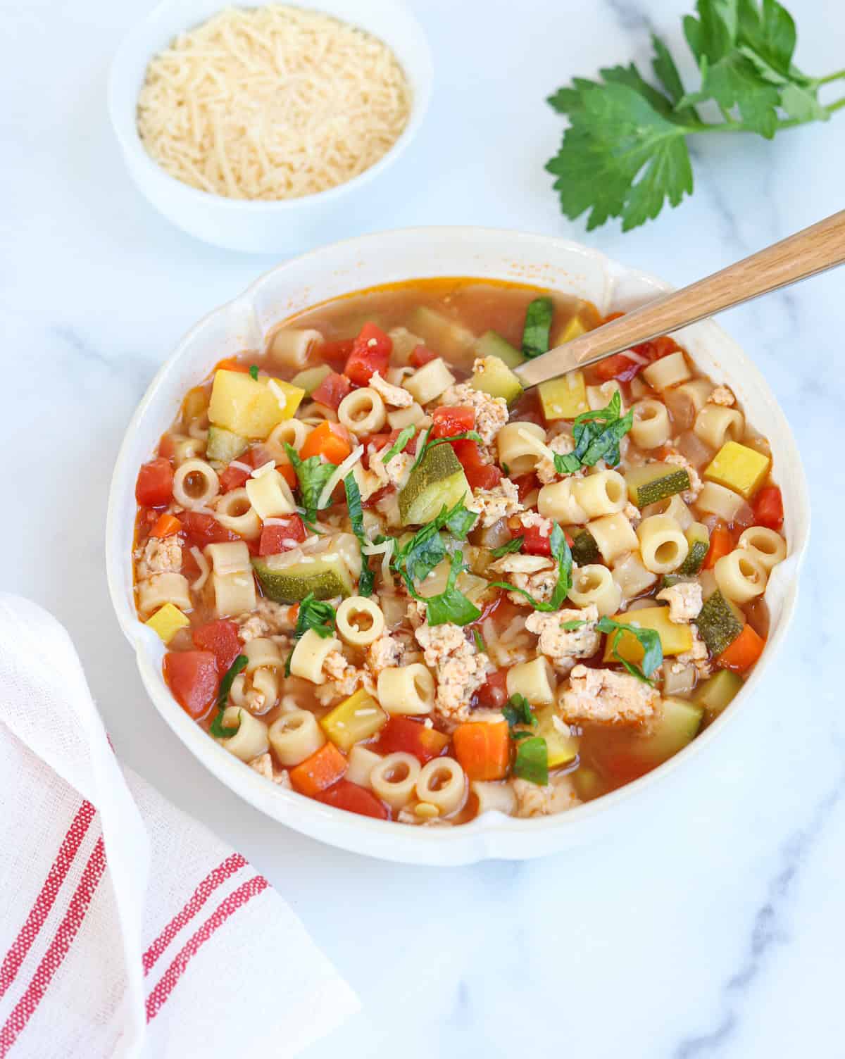 A bowl of vegetable and pasta soup with small pasta, chopped vegetables, and herbs, topped with fresh basil. A wooden spoon, a dish of grated cheese, a bunch of parsley, and a striped towel are nearby on a white surface.
