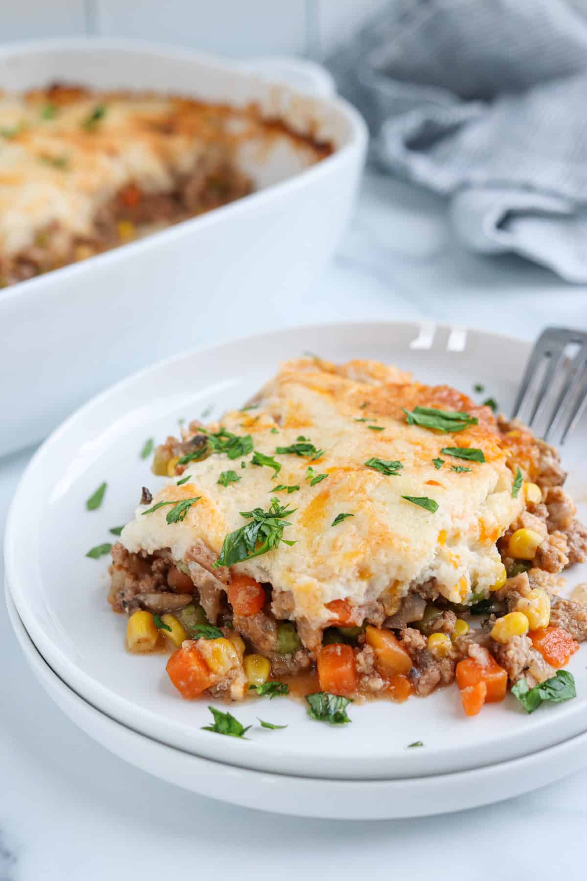 A serving of shepherd’s pie on a white plate, showing layers of ground meat, mixed vegetables, and a golden, cheesy mashed potato topping, garnished with chopped fresh herbs; casserole dish in background.
