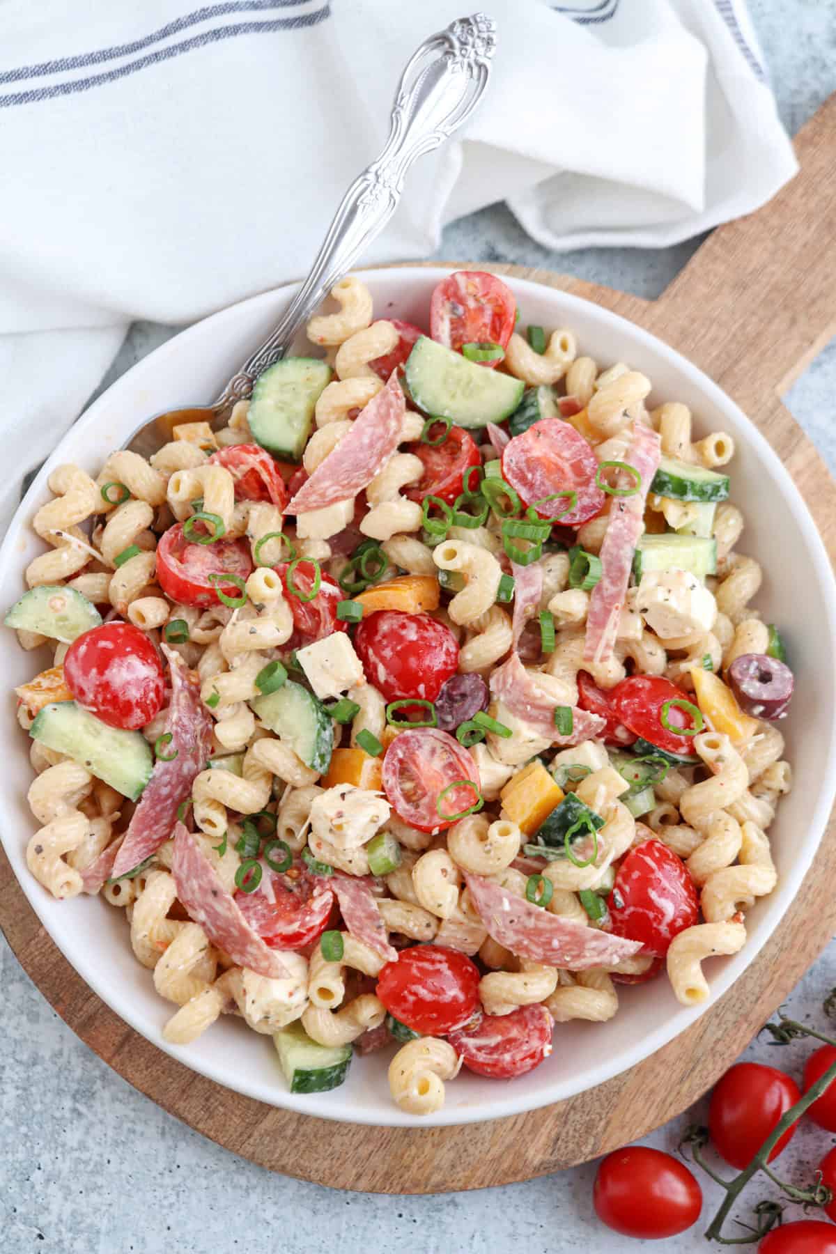 A bowl of pasta salad with spiral noodles, cherry tomatoes, cucumber, salami, cheese cubes, olives, and chopped green onions, placed on a wooden board with a fork and a white napkin nearby.