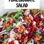 A close-up of a festive pomegranate feta salad with arugula, fresh pomegranate seeds, walnuts, red onion slices, and creamy feta cheese in a white bowl. Text overlay reads Easy Holiday Pomegranate Salad.
