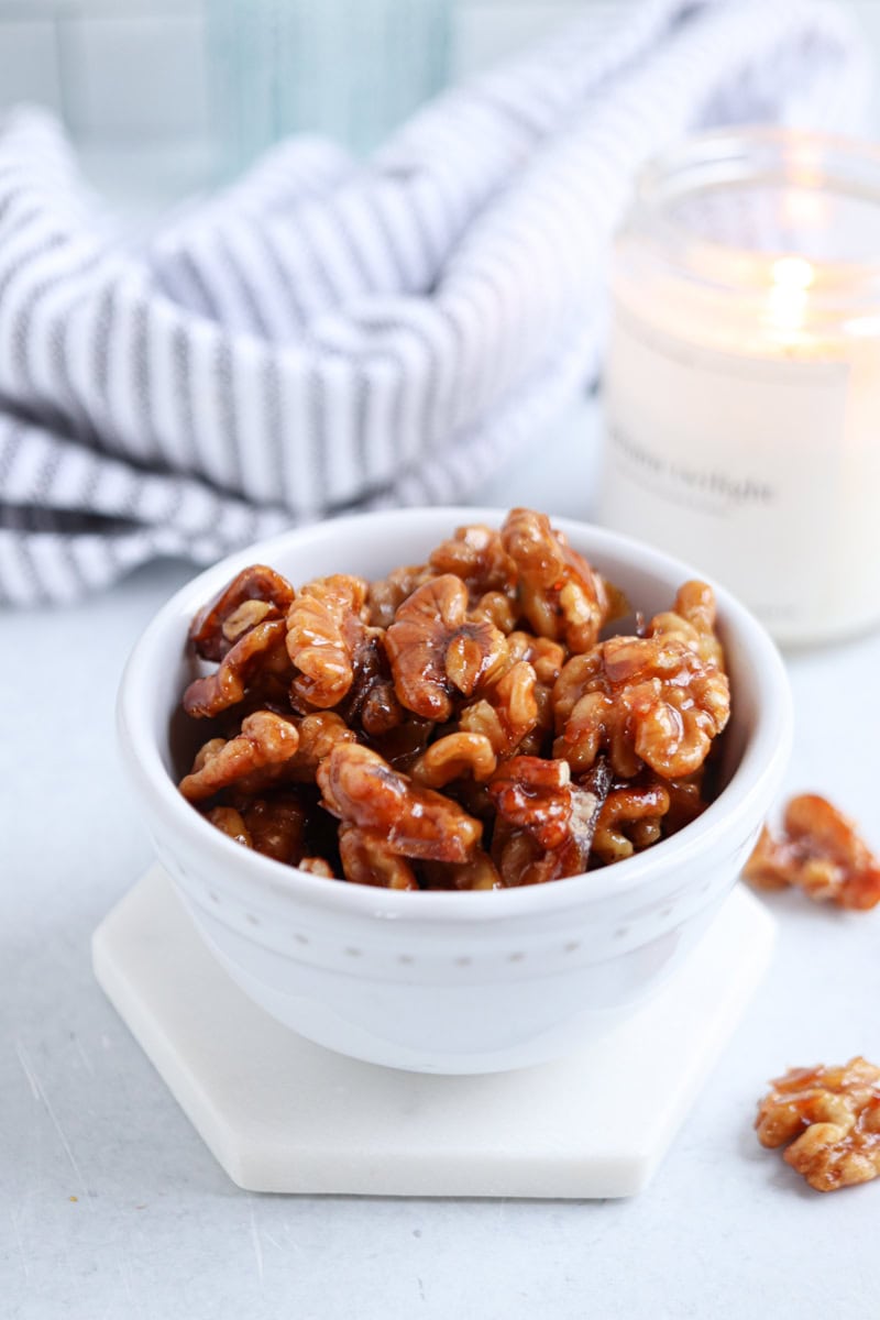 A white bowl filled with glazed walnuts sits on a white coaster, beside a lit candle and a striped cloth—perfect alongside a hearty chicken potato broccoli casserole.