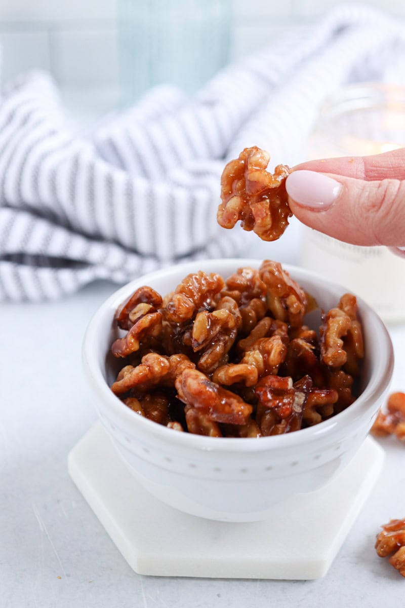 A hand with light pink nails holds a glazed candied walnut above a white bowl filled with more candied walnuts, set beside a striped cloth, candle, and a comforting chicken potato broccoli casserole in the blurred background.
