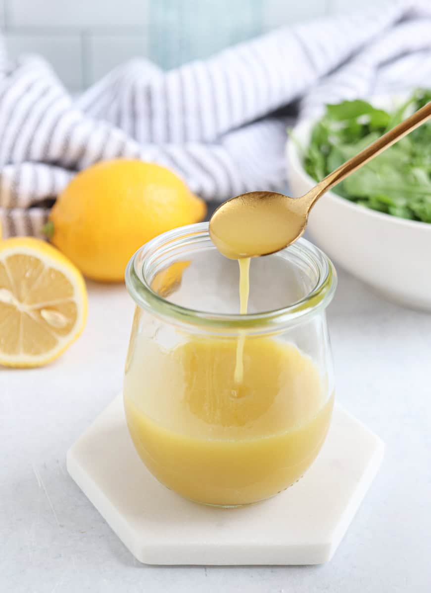 A jar of creamy yellow salad dressing sits on a white coaster, with a gold spoon dripping dressing above it—perfect for drizzling over chicken potato broccoli casserole. A sliced lemon, whole lemon, striped towel, and leafy greens are in the background.