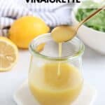A jar of honey lemon vinaigrette with a spoon dripping dressing sits on a white coaster. In the background, a sliced lemon, whole lemon, and fresh greens surround the scene. Text above reads honey lemon vinaigrette.