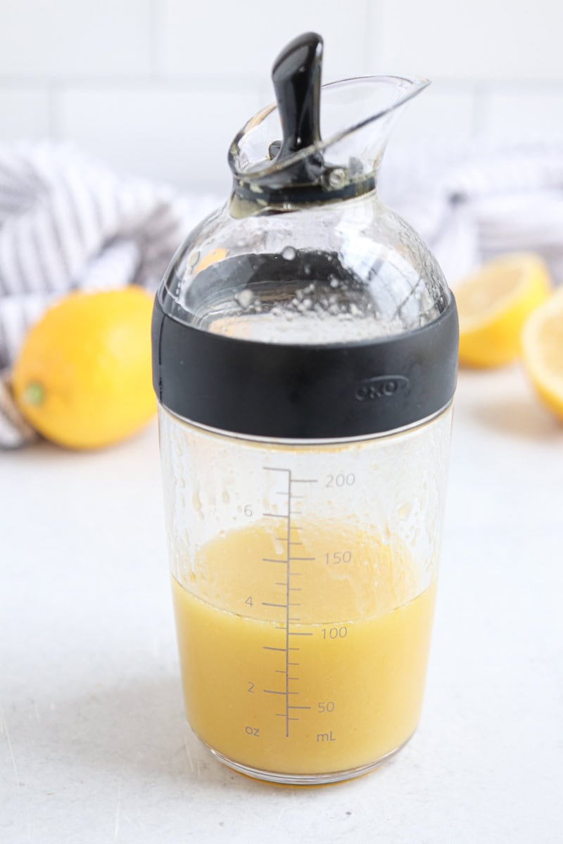 A glass measuring container with a black lid, filled with yellow honey lemon vinaigrette, sits on a white surface. In the background, there are lemons and a striped towel.
