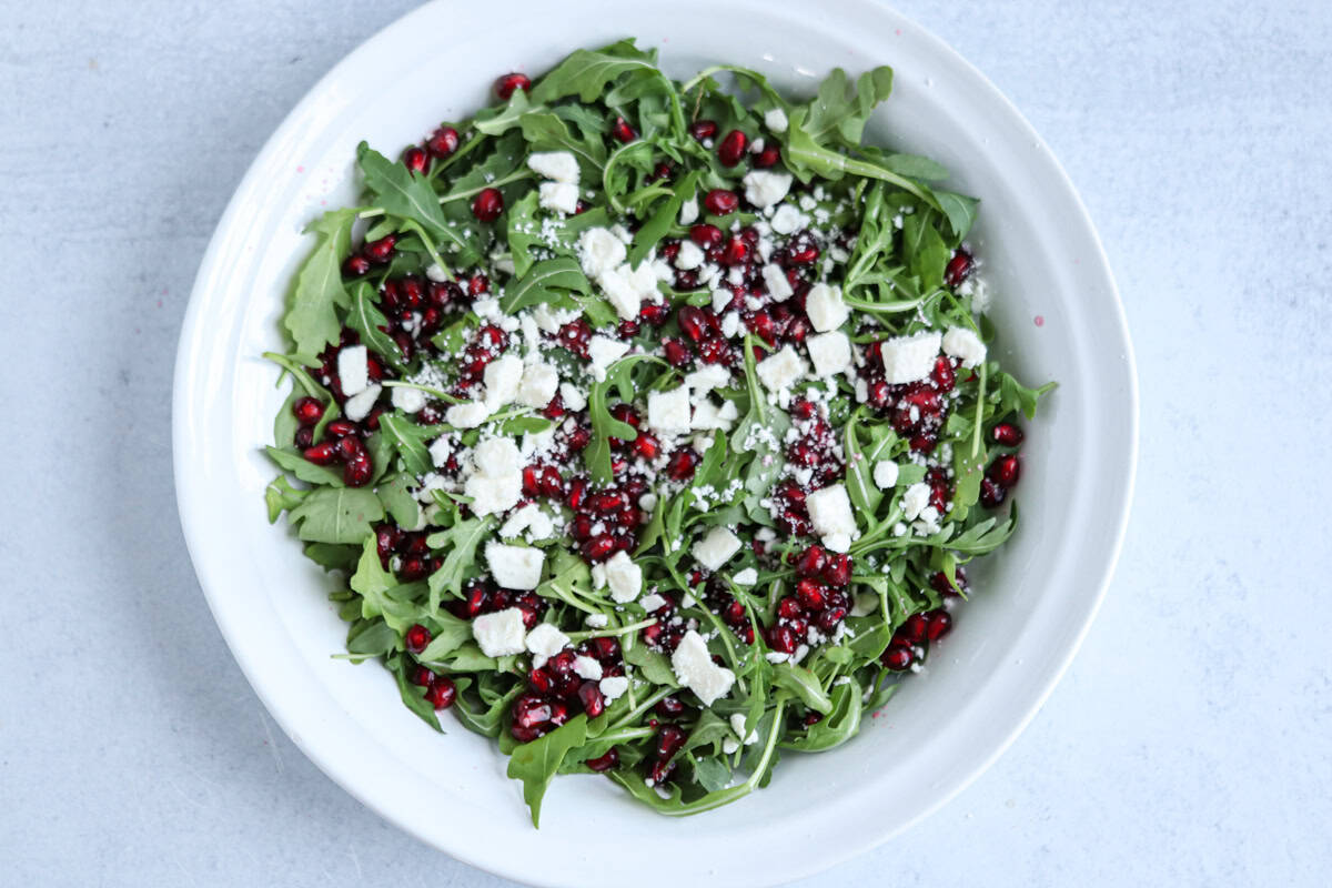 A white bowl filled with arugula leaves, topped with crumbled feta cheese and pomegranate seeds, sits beside a hearty chicken potato broccoli casserole on a light gray surface.