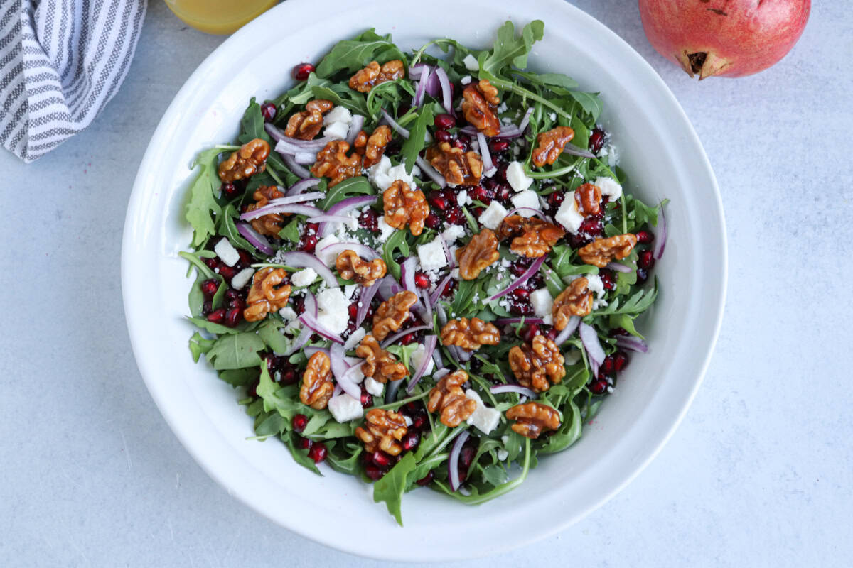 A white bowl filled with arugula, walnuts, pomegranate seeds, feta cheese, and thinly sliced red onions sits on a light surface beside a pomegranate and a striped towel—an elegant contrast to heartier dishes like chicken potato broccoli casserole.