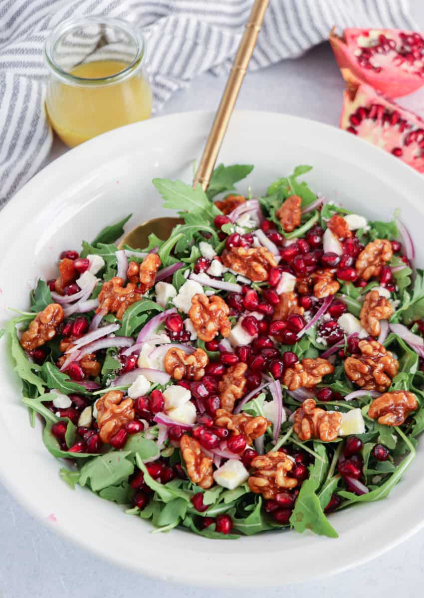 A white bowl filled with arugula salad topped with walnuts, pomegranate seeds, red onion slices, and feta cheese sits beside a gold fork, a small jar of dressing, and a pomegranate—perfect as a side for chicken potato broccoli casserole.