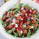 A white bowl filled with arugula, pomegranate seeds, red onion slices, feta cheese crumbles, and walnuts sits beside a serving of chicken potato broccoli casserole, with a gold spoon resting inside.
