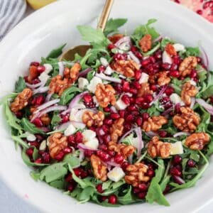 A white bowl filled with arugula, pomegranate seeds, red onion slices, feta cheese crumbles, and walnuts sits beside a serving of chicken potato broccoli casserole, with a gold spoon resting inside.