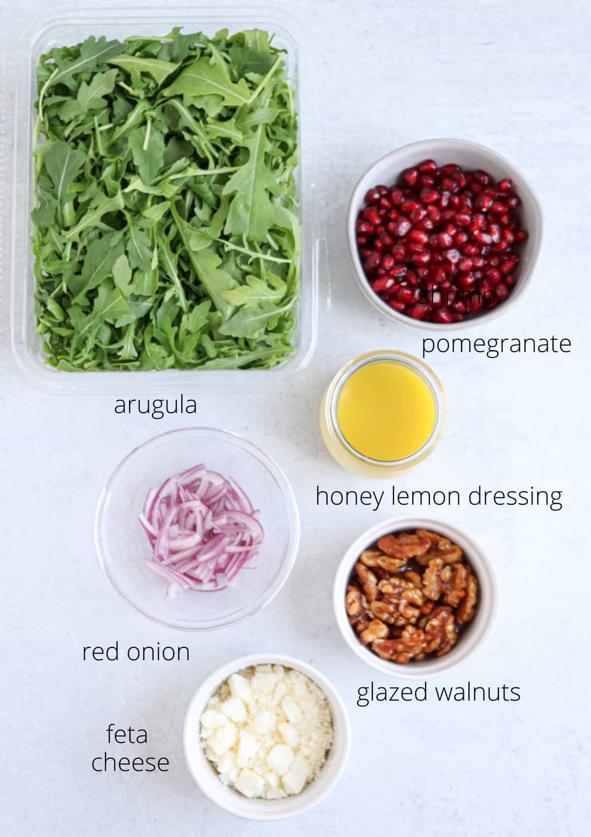 A flat lay of salad ingredients: a container of arugula, a bowl of pomegranate seeds, honey lemon dressing, sliced red onion, glazed walnuts, and crumbled feta cheese—perfect as a side for chicken potato broccoli casserole. Each item is clearly labeled.