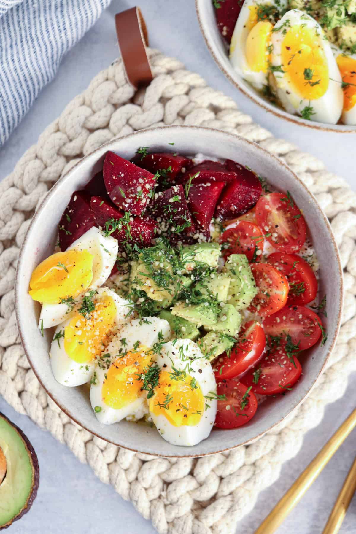 A bowl filled with sliced hard-boiled eggs, avocado, cherry tomatoes, and beetroot, garnished with herbs, sits on a textured placemat with part of an avocado and another bowl nearby.