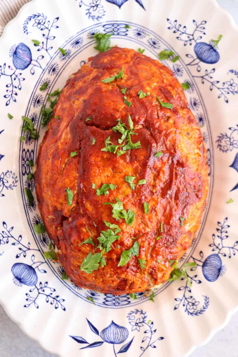 A large, oval-shaped meatloaf topped with tomato sauce and sprinkled with chopped fresh parsley sits on a decorative white and blue floral platter, next to a serving of creamy chicken potato broccoli casserole.