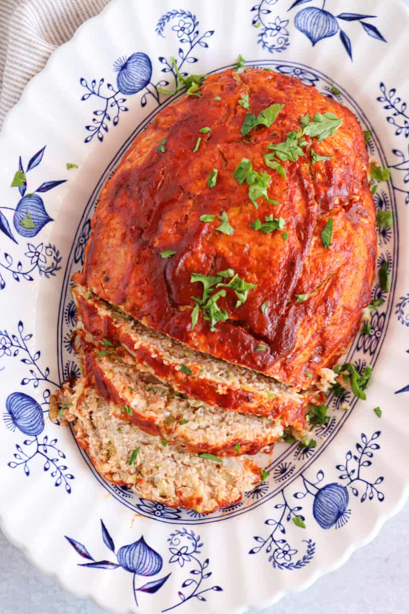 A baked meatloaf topped with tomato sauce and chopped parsley is served on a white and blue floral platter beside a hearty chicken potato broccoli casserole. Several slices are cut, revealing a moist, seasoned interior.