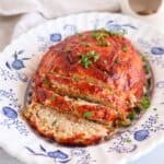 A sliced meatloaf topped with tomato sauce and garnished with chopped parsley is served on a decorative white and blue plate, beside a creamy chicken potato broccoli casserole for a hearty meal.