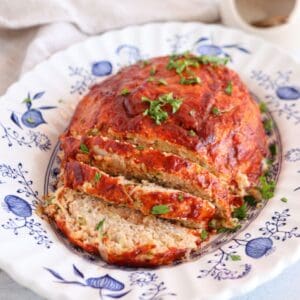 A sliced meatloaf topped with tomato sauce and garnished with chopped parsley is served on a decorative white and blue plate, beside a creamy chicken potato broccoli casserole for a hearty meal.